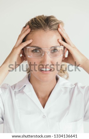 Scientist wearing a lab coat and protective glasses looks stressed, burnt out or has a headache.