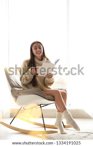 Young woman with cup of coffee reading book near window at home