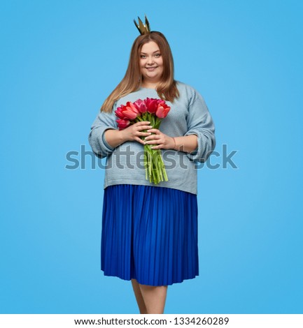 Pretty plus size female in paper crown smiling and looking at camera while holding bunch of bright tulips and standing on blue background