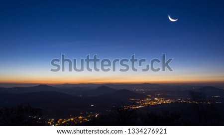 Landscape before sunrise with morning fog and glowing towns in the valleys. Glowing piece of moon high on the sky.