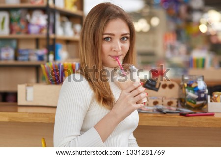 Photo of attractive female drinks milk cocktail with straw, poses in bar, wears white jumper, looks directly at camera, enjoys its taste, has spare time after work or classes. People and lifestyle