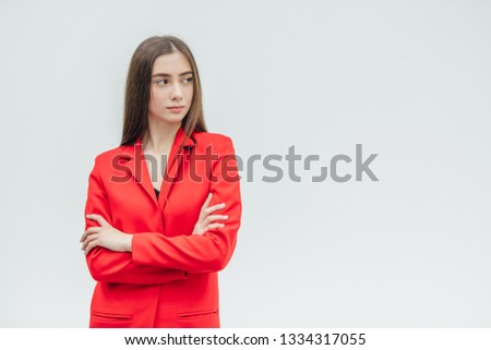 Young pretty, serious girl on a gray background. Standing with a hand on hand. Dressed in a red jacket. With black long hair.