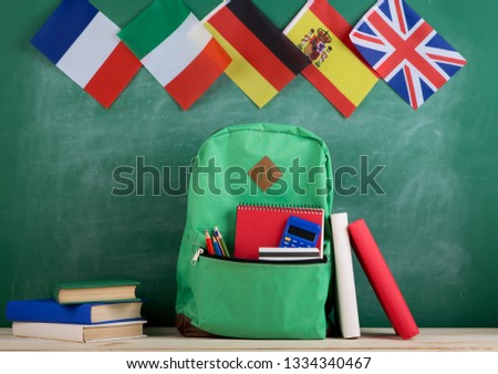 Learning languages concept - green backpack, flags of Spain, France, Great Britain and other countries, books and school supplies on the background of the blackboard