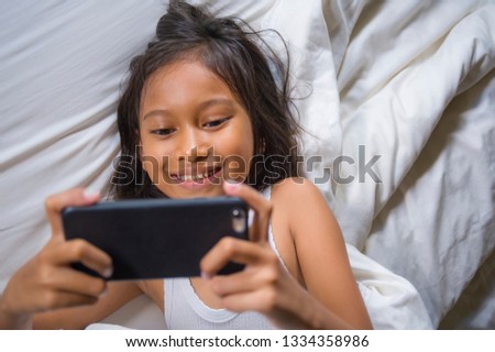 lifestyle portrait of sweet happy and beautiful 7 years old child having fun playing internet game with mobile phone lying on bed cheerful and excited in kid and domestic technology concept