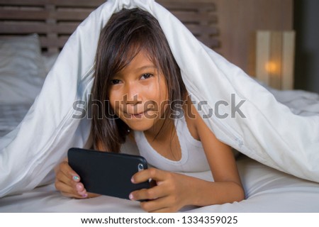 lifestyle portrait of sweet happy and beautiful 7 years old child having fun playing internet game with mobile phone lying on bed cheerful and excited in kid and domestic technology concept