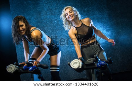 Two attractive young sporty women riding exercise bikes during cycling class indoors