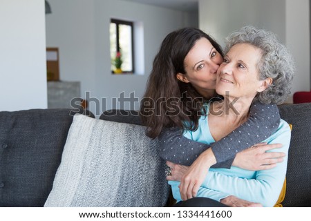 Pretty woman kissing senior mother and hugging her from back. Smiling elderly lady spending time with young daughter. Bonding concept