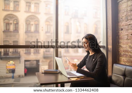 Attractive woman successful commercial real estate analyst checks the status of the account on smartphone while sitting with laptop computer in coffee shop near window with copy space. Online banking  Royalty-Free Stock Photo #1334462408