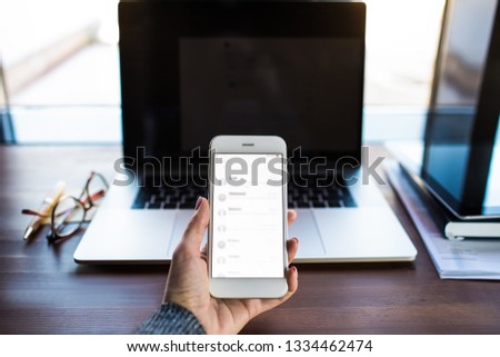 Closely woman blogger holding mobile phone with blurred text messages on the screen against open pc laptop computer and working tools on workstation. Hipster girl using apps on cellphone