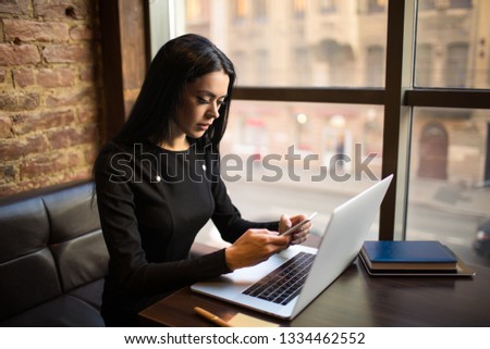 Successful business woman using messenger on mobile phone during training course on laptop computer, sitting in restaurant interior. Serious female magazine editor received notifications on cellphone 