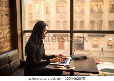 Prosperous woman lawyer using application on pc laptop computer while sitting in restaurant near window with copy space in evening time.Female having online webinar on notebook gadget, resting in cafe
