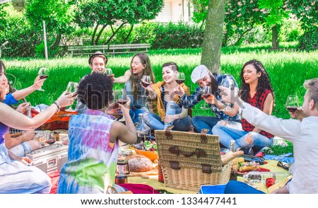 friends drinking wine raising glasses toasting at a garden picnic on a summer day lunch. 10 people get together for picnic lunch in a green park.
