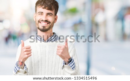 Young handsome man wearing winter sweater over isolated background success sign doing positive gesture with hand, thumbs up smiling and happy. Looking at the camera with cheerful expression, winner