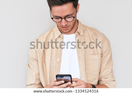 Young man wearing white t-shirt and beige shirt, round trendy eyewear standing isolated on white wall, looking at screen of his cellphone reading messages. Handsome male browsing on smart phone