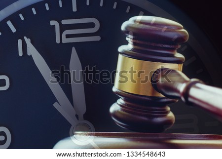 Wood and brass gavel standing upright on its base with a clock dial showing five to twelve behind in a law enforcement or auction concept Royalty-Free Stock Photo #1334548643