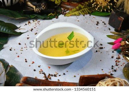 Bone broth made from chicken, served in a white bowl, with green herbs, and flowers in the background - Image