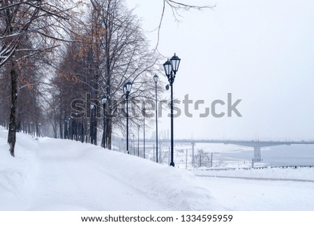 winter snowy alley with vintage lanterns in a city park on the high bank of the river, a concrete road bridge is visible in the distance