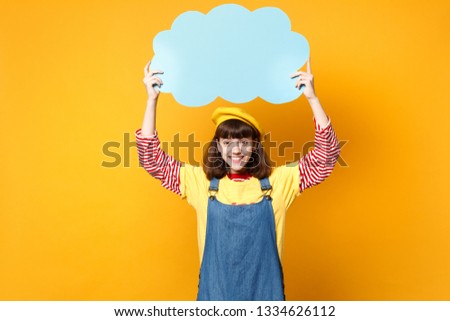 Smiling girl teenager in french beret, denim sundress hold blue empty blank Say cloud, speech bubble isolated on yellow wall background. People sincere emotions, lifestyle concept. Mock up copy space