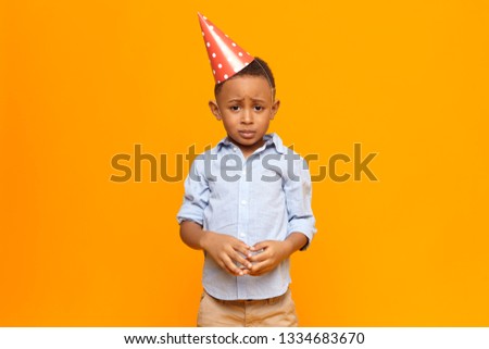 Picture of frustrated unhappy Afro American little boy wearing red cone hat having mournful upset facial expression, making nervous gesture after being told by his father for misbehavior at party