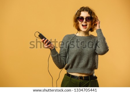 Modern happy millennial teenage girl with sunglasses, headphones smiling showing peace gesture over yellow background, vibrant colors