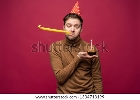 Picture of unhappy stressed young man with birthday cupcake having unhappy look, feeling tired and worn out with birthday party preparations, standing in studio