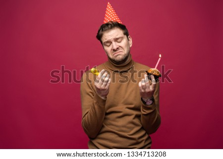Picture of unhappy stressed young man with birthday cupcake having unhappy look, feeling tired and worn out with birthday party preparations, standing in studio