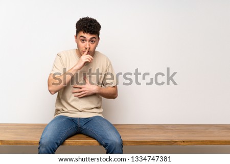 Young man sitting on table showing a sign of silence gesture putting finger in mouth