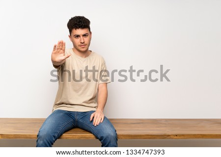 Young man sitting on table making stop gesture