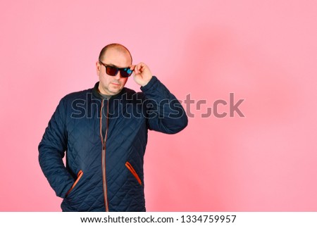 A man in a blue jacket and sunglasses on a pink background in the studio.