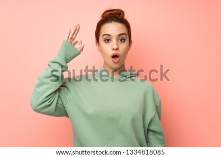 Young redhead woman with sweatshirt surprised and showing ok sign