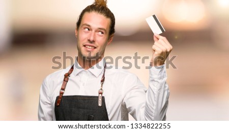 Barber man in an apron holding a credit card and thinking in a barber shop