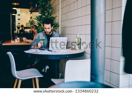 Serious hipster guy holding credit card checking information for making transaction on laptop computer in cafe, pensive male freelancer using online banking for shopping and paying for items