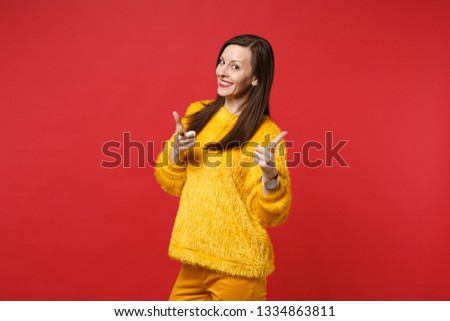 Portrait of smiling young woman in yellow fur sweater pointing index fingers on camera isolated on bright red wall background in studio. People sincere emotions, lifestyle concept. Mock up copy space