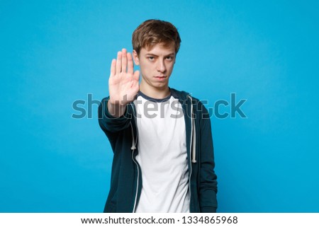 Portrait of serious young man in casual clothes showing stop gesture with palm isolated on blue background in studio. People sincere emotions, lifestyle concept. Mock up copy space. Advertising area