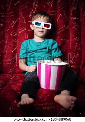 Children's cinema: a little boy watches a movie in 3D glasses on a red sofa and eats popcorn.