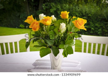 Yellow and white roses in white vase on a table with a white table cloth and two white chairs in the background set in a garden