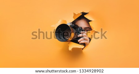 A teenage girl is holding a black camera with a telephoto lens that looks out through a torn hole in yellow paper. Concept of paparazzi, espionage, yellow press. Royalty-Free Stock Photo #1334928902