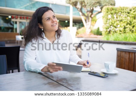 Dreamy woman making notes, working and using tablet in cafe. Young lady wearing white blouse and sitting at table with outdoor cafe interior in background. Freelancer workplace concept.