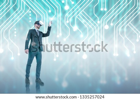 Young businessman with beard wearing dark suit and VR headset working with circuit interface over gray background. Toned image double exposure