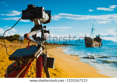 Professional camera on tripod taking picture film video from coastline with rusty shipwreck