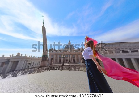 Pretty girl posing with red pink scarf g in front of St. Peter's Basilica, the Vatican City in Rome. Wide angle. Woman wears white top and long blue skirt.
