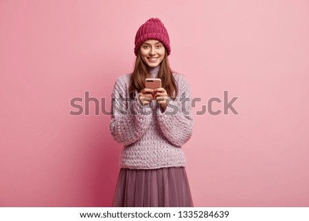 Image of happy positive European female holds mobile phone in hands, waits for order and looks at device screen, plays smartphone game, wears hat, knitted jumper and skirt, isolated on pink wall