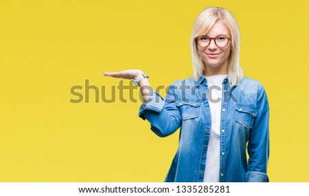 Young beautiful blonde woman wearing glasses over isolated background smiling cheerful presenting and pointing with palm of hand looking at the camera.