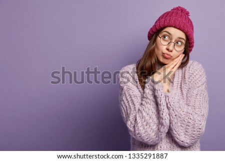 Photo of sad young Caucasian woman feels lonely, focused aside with unhappy thoughtful expression, folds lips, leans on pressed palm together, wears hat and sweater, isolated on purple wall.