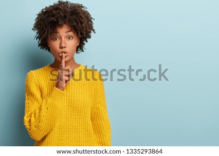 Studio shot of quiet dark skinned lady keeps fore finger over lips, makes silence gesture, gossips with friend, wears yellow sweater, says hush, isolated over blue studio wall with blank space Royalty-Free Stock Photo #1335293864