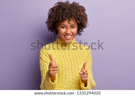 I choose you. Smiling delighted young female with Afro haircut, points with two fingers, makes choice, wears yellow jumper, isolated over purple background, shows her decision. Join me, please Royalty-Free Stock Photo #1335294020