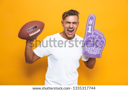 Image of cheerful man 30s in white t-shirt holding rugby ball and number one fan hand glove with finger raised while standing isolated over yellow background