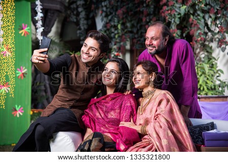 Indian family taking selfie picture using smartphone while wearing traditional festival cloths on diwali/wedding ceremony and sitting on sofa / couch