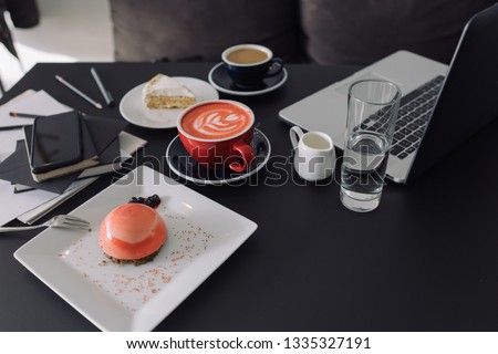 Open laptop, cups of coffee, dessert, stationery on black background. Freelancer work concept. Space for text, sunrise light.
