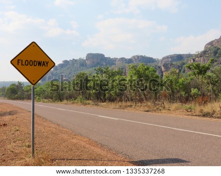Floodway sign next to the road surrounded by a dry landscape with green trees and mountains. Located in Australia. 